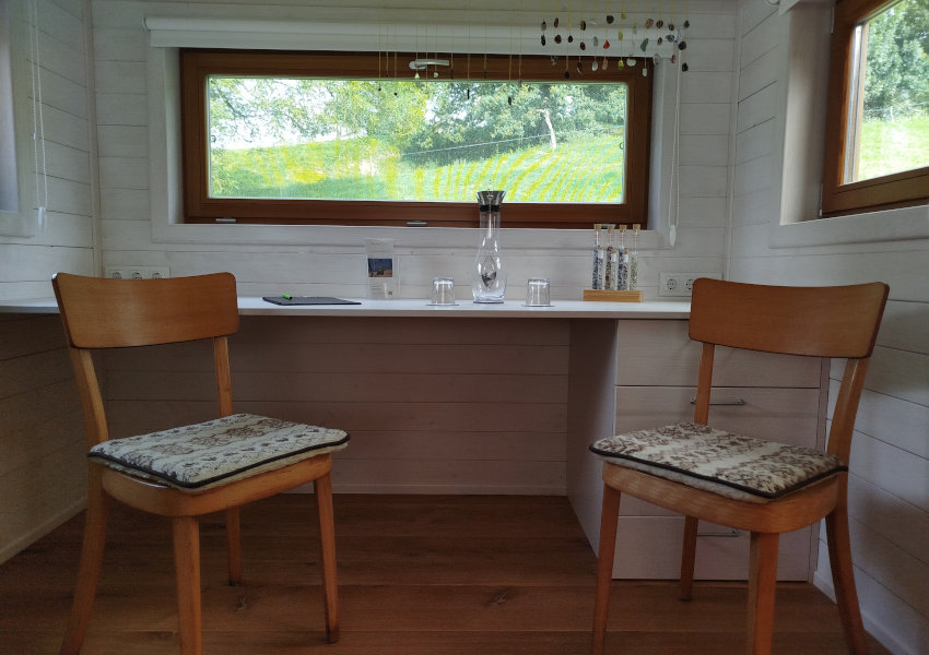 Blick auf die Sitzecke mit den zwei St&uuml;hlen und dem Tisch. Auf dem Tisch steht eine Glaskaraffe mit zwei Trinkgl&auml;sern. Im Hintergrund sieht man ein Fenster mit Blick auf eine gr&uuml;ne Wiese.